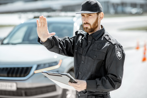 Police officer holding a tablet and raising his hand to signal a stop beside a parked car.