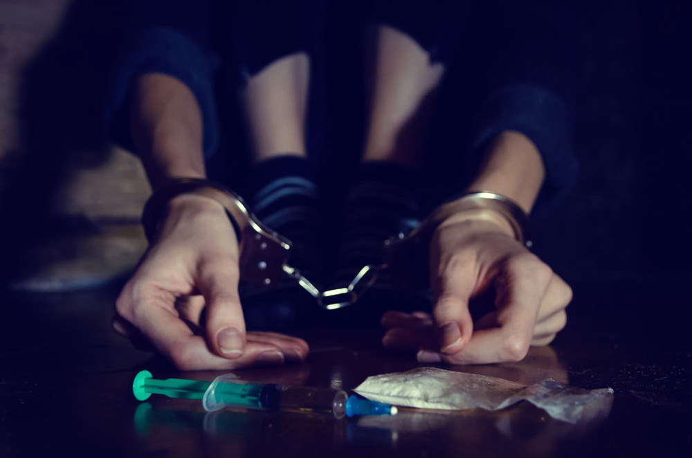 Handcuffed hands resting on a dark table next to small bags of drugs.