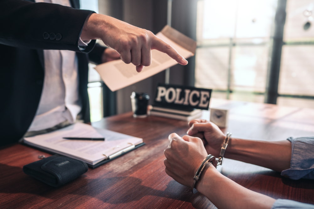 Handcuffed person seated at a desk across from an officer taking notes beside a police nameplate.