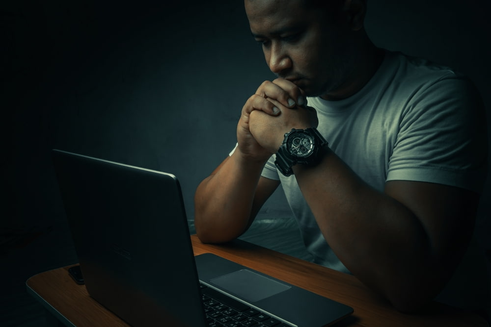 A man sitting at a laptop with his hands clasped in thought in a dimly lit room.