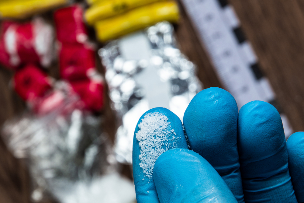 Close-up of blue gloved fingers holding a small amount of white crystalline powder, with other items blurred in the background.