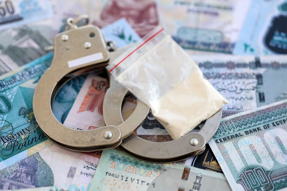 Metal handcuffs and a plastic bag containing white powder displayed on assorted international banknotes.